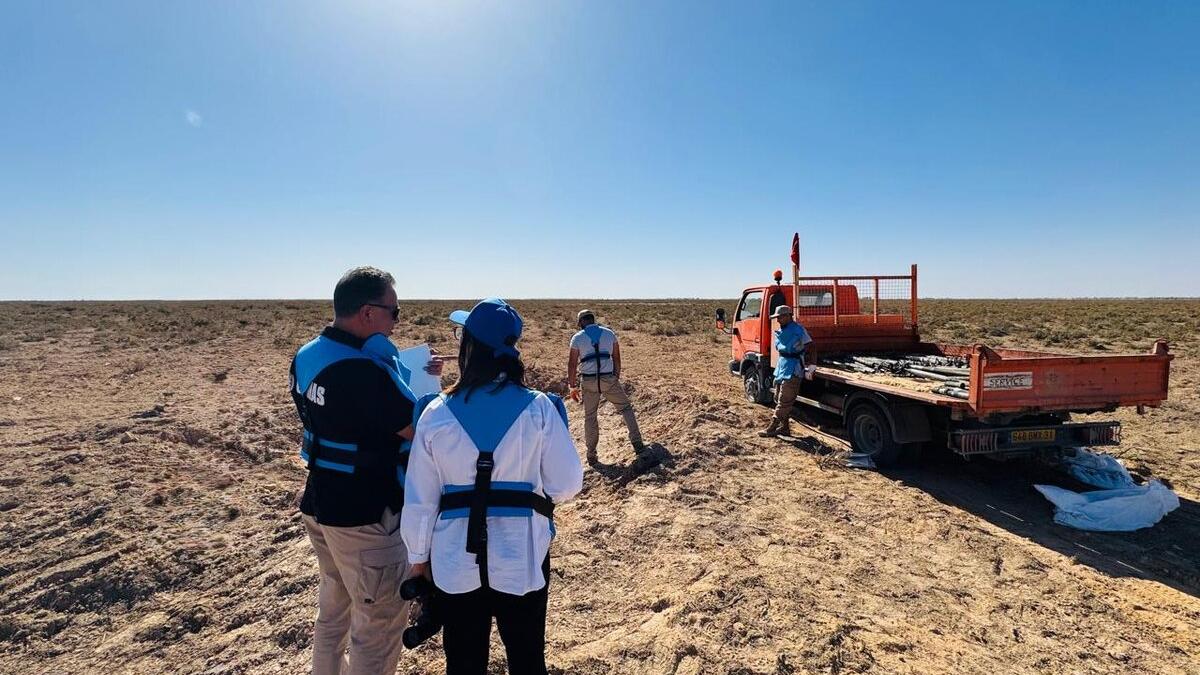 A wider shot of the operation site under bright sunlight. In the foreground, two individuals (a man and a woman) are seen from behind; the man is wearing a black vest with "UNMAS" lettering, and the woman is wearing a white shirt with blue protective gear. They appear to be supervising or documenting the work. In the background, the orange truck is parked with its tailgate down, and the bed is now loaded with several of the rusted munitions. The landscape extends into a flat, horizon-less desert.
