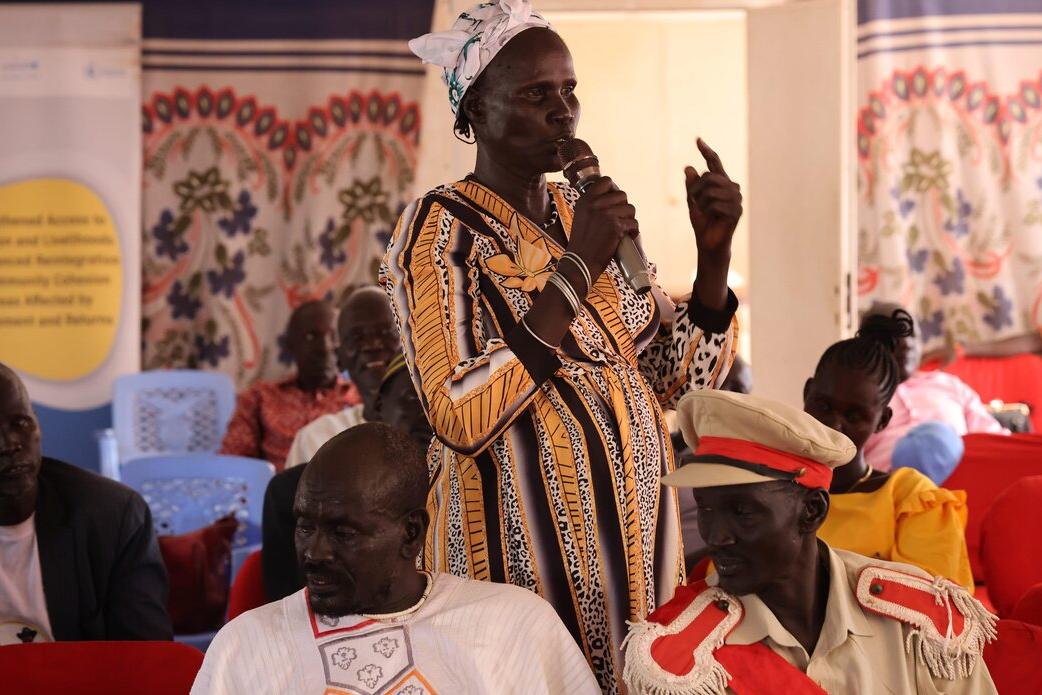 Person speaking into a microphone while standing among seated participants in a hall decorated with patterned fabric and banners.