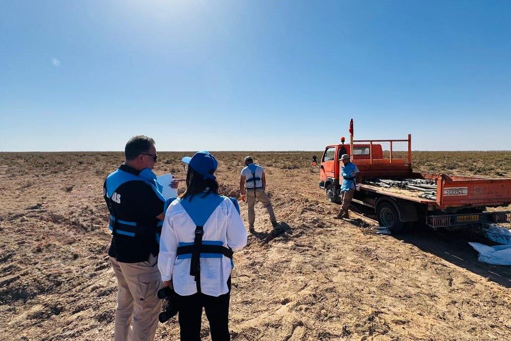 A wider shot of the operation site under bright sunlight. In the foreground, two individuals (a man and a woman) are seen from behind; the man is wearing a black vest with "UNMAS" lettering, and the woman is wearing a white shirt with blue protective gear. They appear to be supervising or documenting the work. In the background, the orange truck is parked with its tailgate down, and the bed is now loaded with several of the rusted munitions. The landscape extends into a flat, horizon-less desert.
