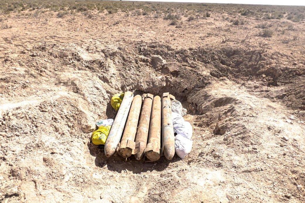 Two field operators are shown in the midst of handling the ordnance. Both men are wearing light grey uniforms, caps, and distinct blue protective body armor vests. They are wearing heavy-duty gloves and are carefully passing a rusted projectile from one person standing on the ground to another standing on the bed of an orange utility truck. The truck features a cage-like barrier behind the cab and a red flag attached to a pole, serving as a safety warning signal.