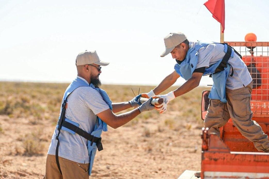 A high-angle shot looking down into a shallow, dug-out pit in dry, rocky desert soil. Inside the pit, approximately five to six large, rusted cylindrical munitions (likely large-caliber artillery shells or rockets) are carefully laid out in a row. They are resting on top of white and yellow sandbags, likely placed there to stabilize the ordnance or prepare it for controlled demolition or safe transport. The surrounding area is barren desert scrubland.